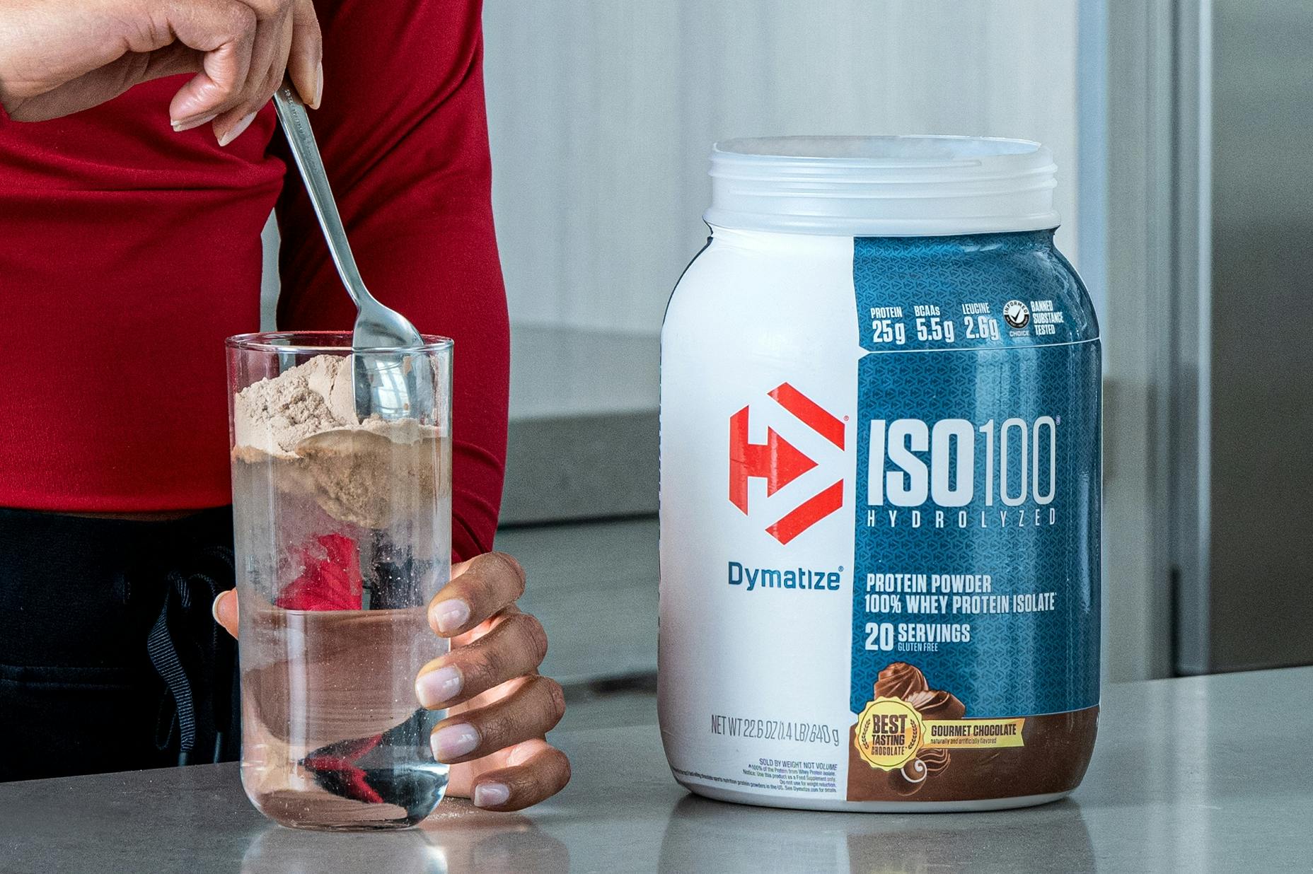 A person in a kitchen, stirring Dymatize ISO100 powder into a glass.